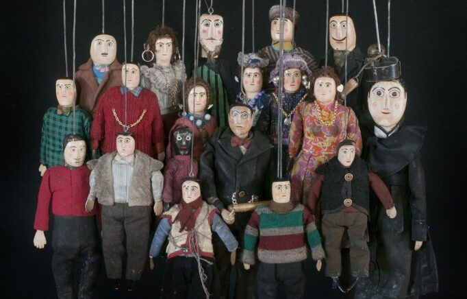 Photographie ressemblant à une photo de famille de multiples marionnettes différentes représentant des hommes et des femmes tenus par une tringle sortant de la tête.