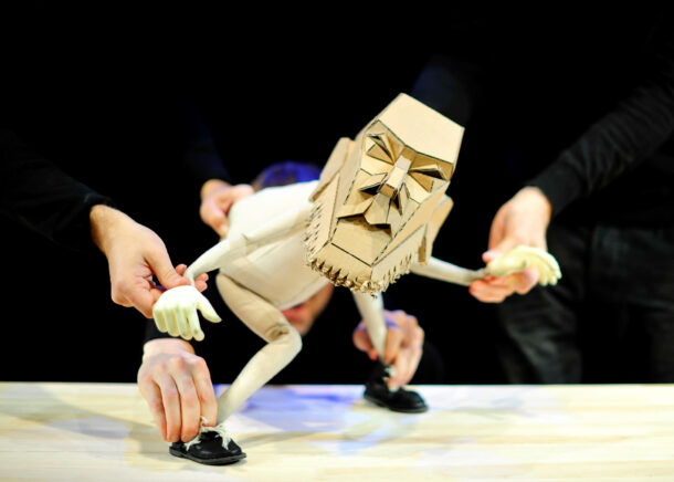 Une marionnette au visage en carton, posée sur une table, est manipulée par trois marionnettistes