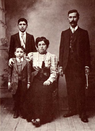 Photographie de famille en noir et blanc où l'on voit un homme en costume sur la droite, le regard pétillant, une femme au centre en jupe et chemise à gros carreaux avec un visage strict, et sur le côté gauche deux jeunes garçons en costume.