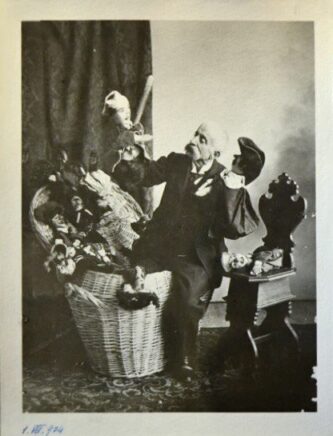 Photographie d'un homme à moustache blanche ayant une jambe dans un gros panier en osier rempli de marionnettes et l'autre à l'extérieur. Il tient une marionnette de Guignol dans chaque main.