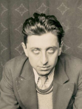 Un homme portant un pull et un costume, les cheveux mi-longs ébourrifés, regardant avec un insistance face à l'objectif.