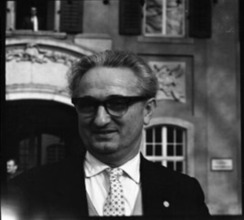 Un homme relativement âgé en costume avec des lunettes devant un théâtre, le visage légèrement souriant et tourné à gauche.