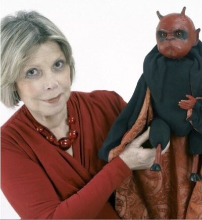 Photographie en couleur d'une femme grisonnante portant vers la caméra une marionnette de diable
