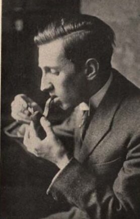 Un homme jeune en costume et avec les cheveux mi-long tourné vers la gauche tentant d'allumer sa pipe.