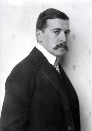 Un homme en costume tourné vers la droite avec des cheveux courts et une grosse moustache.