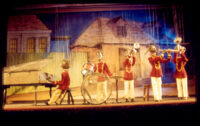 Cinq marionnettes à fils forment un orchestre de jazz en uniformes rouges, devant un décor de maisons anciennes.