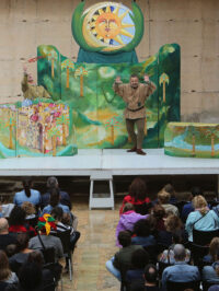 Photographie d'un spectacle où l'on voit le public assis sur des chaises devant la scène où se trouve un acteur en costume médiéval vert entouré d'un décor de jungle.