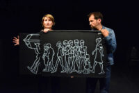 Photographie d'un spectacle où l'on voit un homme et une femme tenir devant eux un tissu où il est dessiné un groupe de personnes marchant derrière un cercueil porté par des hommes.