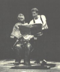 Un homme sur scène avec des bretelles tenant un livre qu'il montre à un mannequin assis sur une chaise.
