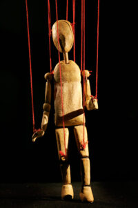 Petite marionnette en bois tenue par des fils rouges représentant un humain sans visage.