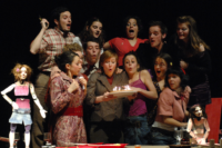 Photographie d'une scène de la pièce où l'on voit 11 personnes et deux marionnettes en rang, entourant une dame qui souffle des bougies sur un gâteau devant la table d'un repas.