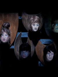 Une photographie de spectacle où l'on voit des sculptures de visage de femme accrochées à un mur via une planche en bois, exhibées comme un trophée.
