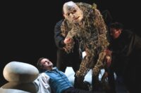 Un homme couché regarde une grande marionnette manipulée par derrière