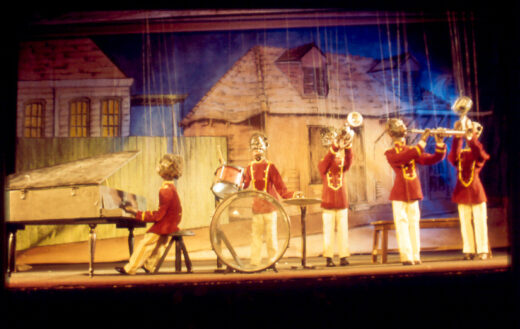 Cinq marionnettes à fils forment un orchestre de jazz en uniformes rouges, devant un décor de maisons anciennes.