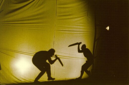 Photographie d'un spectacle de théâtre d'ombre où l'on voit deux personnages courbés avec des couteaux se faisant face.