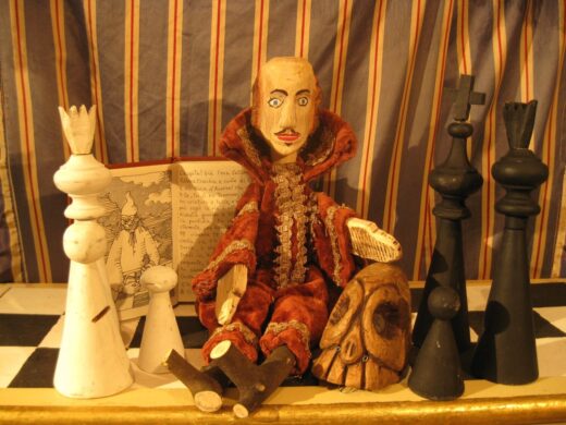 Plusieurs objets sont posés sur un échiquier: quatre pièces du jeu d'échecs, une marionnette représentant Shakespeare, un crâne en bois et un livre ouvert.