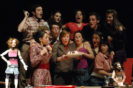 Photographie d'une scène de la pièce où l'on voit 11 personnes et deux marionnettes en rang, entourant une dame qui souffle des bougies sur un gâteau devant la table d'un repas.