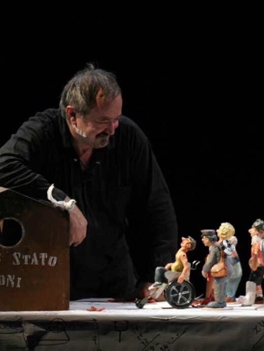 Photographie d'un spectacle où un homme regarde des petites figurines sur une table représentant un homme en fauteuil roulant et différents personnages autour.