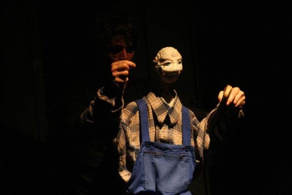 Photographie de spectacle où l'on voit une marionnette habillée en chemise à gros carreaux et salopette bleu d'usine.reaux et salopette bleu