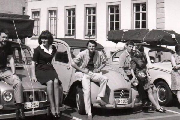 Photographie ancienne montrant des jeunes gens, les femmes en robe et les hommes en pantalons et veste légère, assis sur le capot de plusieurs Citroën 2CV.