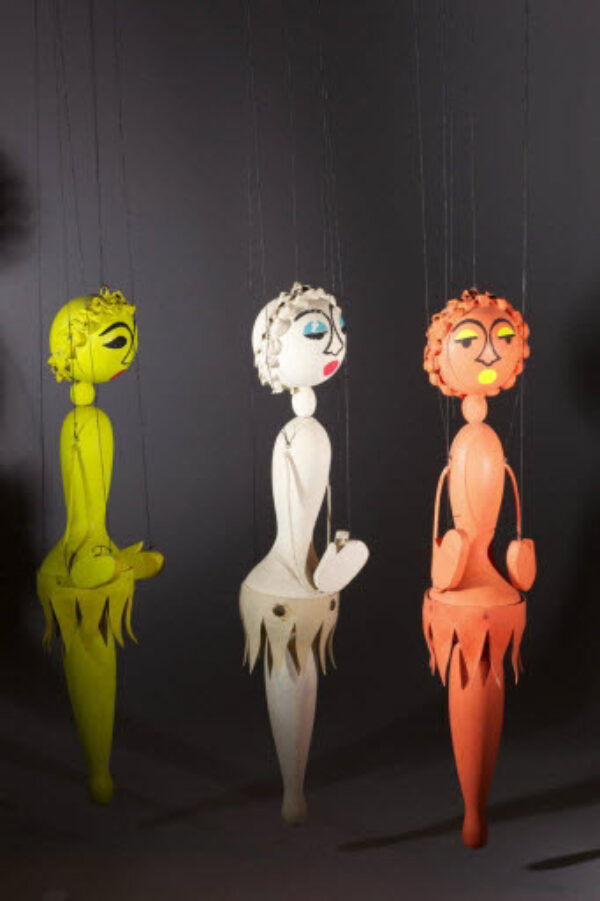 Une photographie de 3 marionnettes à fils de couleur différentes (jaune, rose, blanche) avec un corps filiforme, un visage en forme de tournesol et une courte jupe.