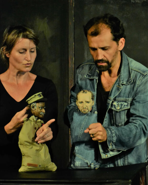 Photographie où l'on voit un homme et une femme manipuler deux petites marionnettes sur une table, l'une des marionnettes a une tenue en jean et l'autre un uniforme d'officier militaire.