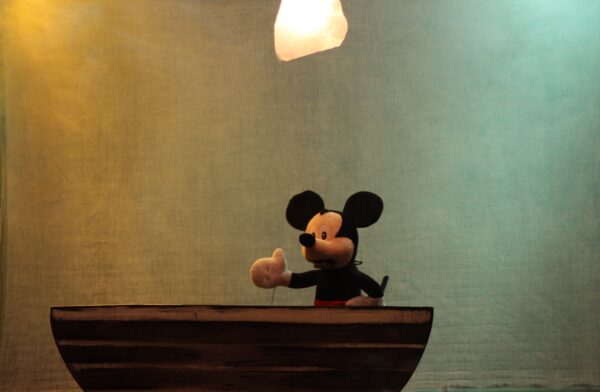 Une marionnette de Mickey Mouse, personnage de Disney, manipulée par des tiges fines sur les mains, assise derrière une table.