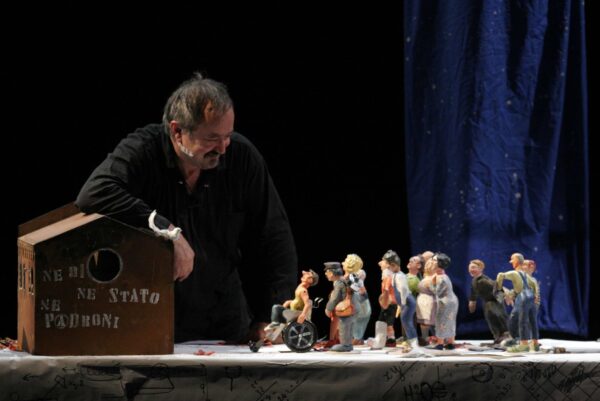 Photographie de spectacle d'un homme accoudé sur une petite maison posée sur une table et regardant une foule de figurines représentant des personnes en tout genre.