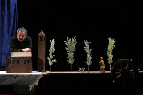 Photographie d'un spectacle représentant un homme accoudé sur une petite maison sur une table. La table comporte, outre la maison, une tour, des arbres et deux figurines d'un chien et d'un homme.