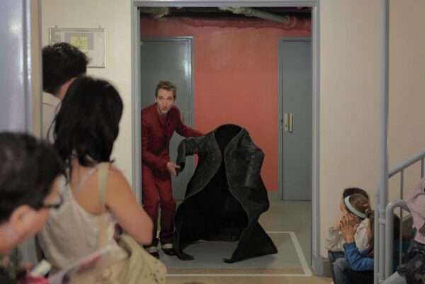 Un homme en costume rouge se tient dans l'encadrement d'une porte en tenant un manteau large qui semble habité. De nombreuses personnes le regardent en nous tournant le dos.